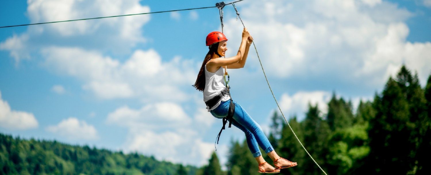 Young woman in casual wearing with red helmet riding on a zip line in the mountains. Active kind of recreation